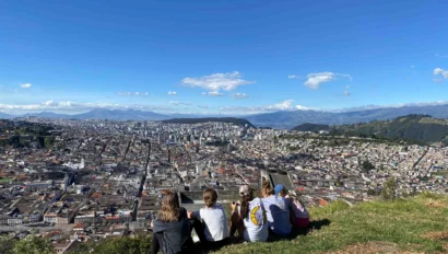 Five people sit on a hillside overlooking a sprawling cityscape under a clear blue sky.