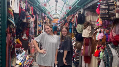 Two people stand in an indoor market aisle with colorful textiles, clothing, and hats displayed on either side.