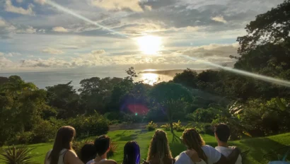 A group of people standing arm in arm on a grassy area, watching the sunset over a distant ocean.