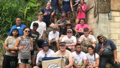 A group of people in casual clothing pose on a muddy slope in front of a building, holding a Georgia Tech banner. Some are wearing gloves and boots, suggesting involvement in a building project.