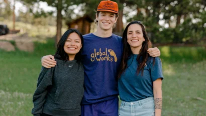 Three people standing closely together outdoors in a grassy area, smiling at the camera. The person in the middle is wearing an orange cap and a blue "global works" T-shirt.