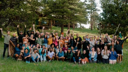 A group of people poses together outdoors on a grassy field with trees and a cabin in the background.