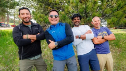 Four men stand outdoors in casual clothing, smiling with arms crossed. They are in front of a tree, surrounded by grass and other trees.