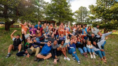 A large group of people posing enthusiastically outdoors on a grassy field, surrounded by trees, under a cloudy blue sky.