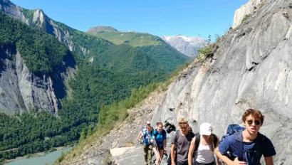 A group of hikers walks along a narrow mountain path with a river and green mountains in the background under a clear blue sky.
