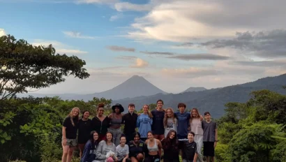 A group of people posing on a grassy area with a distant volcano and cloudy sky in the background.