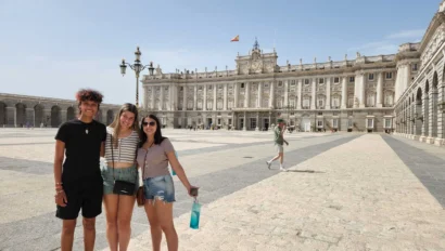Three people stand smiling in front of the Royal Palace of Madrid on a sunny day. A person walks in the background.