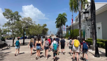 A group of people with backpacks walk down a sunny street lined with palm trees and buildings.