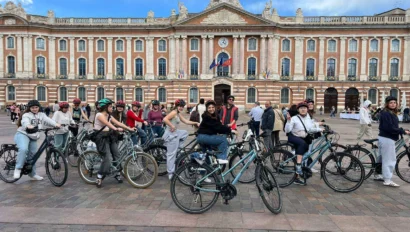 A group of people on bicycles gather in front of a historic building with a clock and flags on a clear day.
