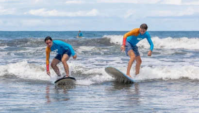 Two people surfing small waves near the shore under a cloudy sky.