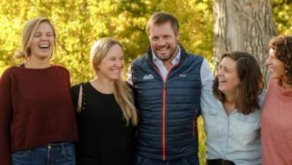 Five people standing outdoors, smiling and laughing in a grassy area with trees and sunlight in the background.