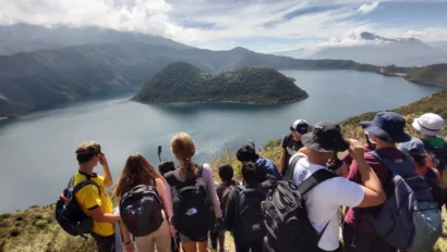 A group of student hikers with backpacks, part of the student travel programs offered by Global Works, stands on a hillside overlooking a large lake and green island, with mountains in the background under a partly cloudy sky.