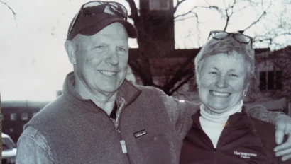 A smiling man and woman with short hair stand outdoors. Both wear sunglasses on their heads and casual vests. A bare tree and building with a clock tower are in the background.