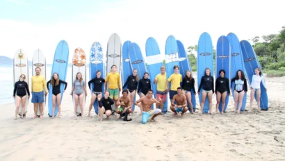 A group of people in swimsuits and rash guards stand on a beach, each holding a surfboard vertically in the sand. Trees and the ocean are visible in the background.