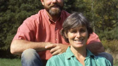 A man and woman pose outdoors on a sunny day, sitting on a rock with trees in the background. The man wears a red shirt, and the woman wears a green shirt.