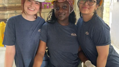 Three student travelers during a school group travel program wearing hard hats, gloves, and matching blue shirts as they pose indoors at a construction site, smiling while working with metal rods and wooden planks.