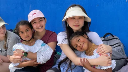 Three girls pose with two young children wearing traditional attire against a blue wall. One girl wears a visor, another a pink hat.