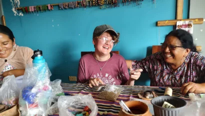 Three people sit at a table with craft supplies, smiling and laughing together. Beaded jewelry hangs on the blue wall behind them.