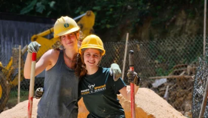 Two people wearing yellow hard hats and holding shovels stand next to each other in a construction area with a pile of sand and a bulldozer in the background.