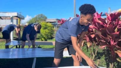 A young person on a service trip for teens uses a paint roller to apply white paint to a large panel outdoors, while others paint similar panels in the background. Bright red plants and blue sky are visible.