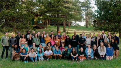 A large group of people posing outdoors on a grass field, with trees and cabins in the background.