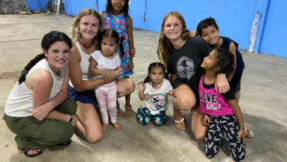 Three young women and five young children pose together, smiling, in a large indoor space with blue walls and a concrete floor—capturing the spirit of teen programs that work with kids.