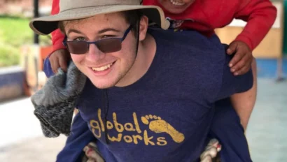 A student wearing sunglasses and a Global Works t-shirt, participating in teen programs that work with kids, gives a piggyback ride to a smiling child outdoors.