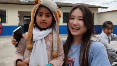 A child wearing a monkey hat stands with a smiling young woman outdoors. Another child is in the background.