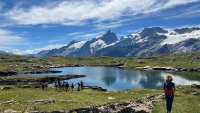 A group of student travelers by a lake with mountains in the background, while one person with a backpack—part of a student travel program—walks toward them under a partly cloudy sky.