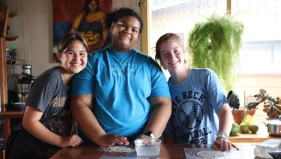 Three people smiling at a table with tortillas, a bowl of water, and plants in the background.