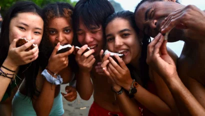Five people stand on a beach, smiling and eating pieces of coconut together.