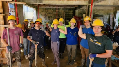 A group of people wearing yellow hard hats and work gloves, posing with tools in a construction site.