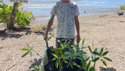 A person stands on a beach, pushing a wheelbarrow full of young plants during a conservation trip. In the background, a sign reads, "Be like. Pick up your trash".
