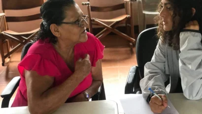 An older woman in a pink top speaks to a younger woman taking notes with a pen and paper at a table indoors during a Spanish immersion program.