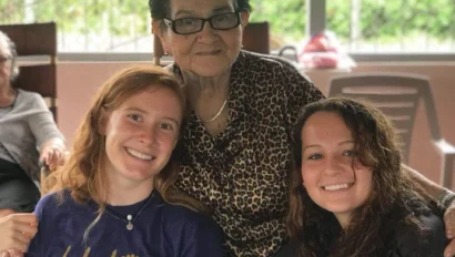 Three women pose together and smile at a table outdoors. The older woman stands behind two younger women, one in a “global works” t-shirt while experiencing their student travel to Costa Rica. Greenery is visible in the background.