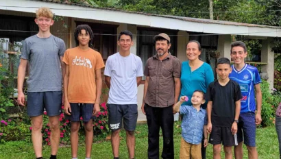 A group of eight people, including adults and children, stand in a row on grass in front of a house with flowers during their Costa Rica homestay; a black and white cat is in the foreground.