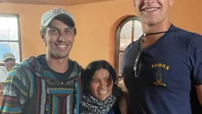 Three people stand indoors smiling at the camera; two young men flank an older woman. The warm-colored walls and natural light evoke the relaxed mood of summer travel in this casual setting.