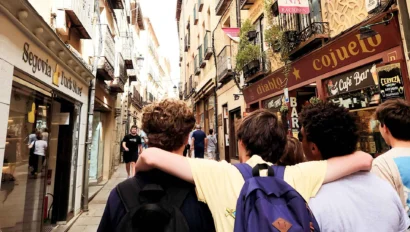 A group of teenagers with backpacks walk down a narrow street lined with shops and balconies in a European town, enjoying their spanish immersion program to spain.