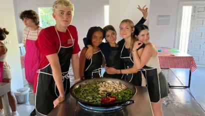Five teenagers wearing aprons stand around a large pan of vegetables in a bright kitchen, posing for a group photo during their spanish immersion program to Spain.