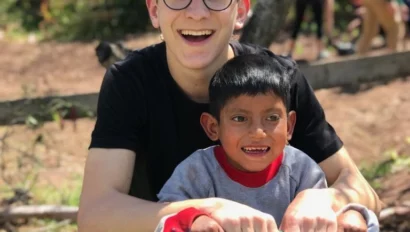 A smiling young adult and a child sit on a seesaw in an outdoor setting with trees and people in the background.
