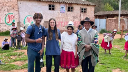 Four people stand together smiling in front of a rustic building, capturing the spirit of Peru student travel, with a group of children and adults in the background in an outdoor rural setting.