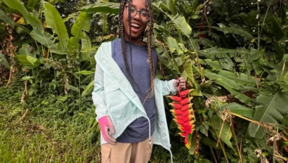 A student traveler standing outdoors in front of tropical plants, smiling and holding a hanging cluster of red and yellow flowers during student travel to Puerto Rico.