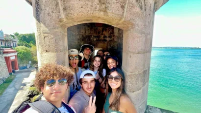 A group of young adults on a student travel to Puerto Rico pose for a photo inside a stone turret at El Morro by the waterfront on a sunny day.