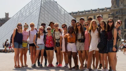 A group of young people on a student trip to France poses for a photo in front of the Louvre Pyramid in Paris on a sunny day.