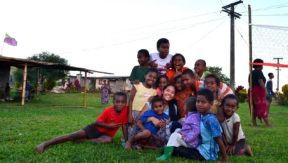 A group of children and an adult, part of a summer service program, sit and kneel together on green grass, smiling at the camera in an outdoor setting with buildings and utility poles in the background.