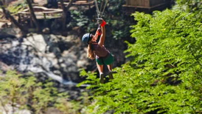 A person wearing a helmet and harness glides on a zip line above a forested area, making this the perfect adventure travel for teens with trees and a river below.