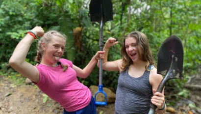 Two young women, part of a Global Works student travel program, pose and flex their arms while holding shovels in a muddy, forested outdoor setting.