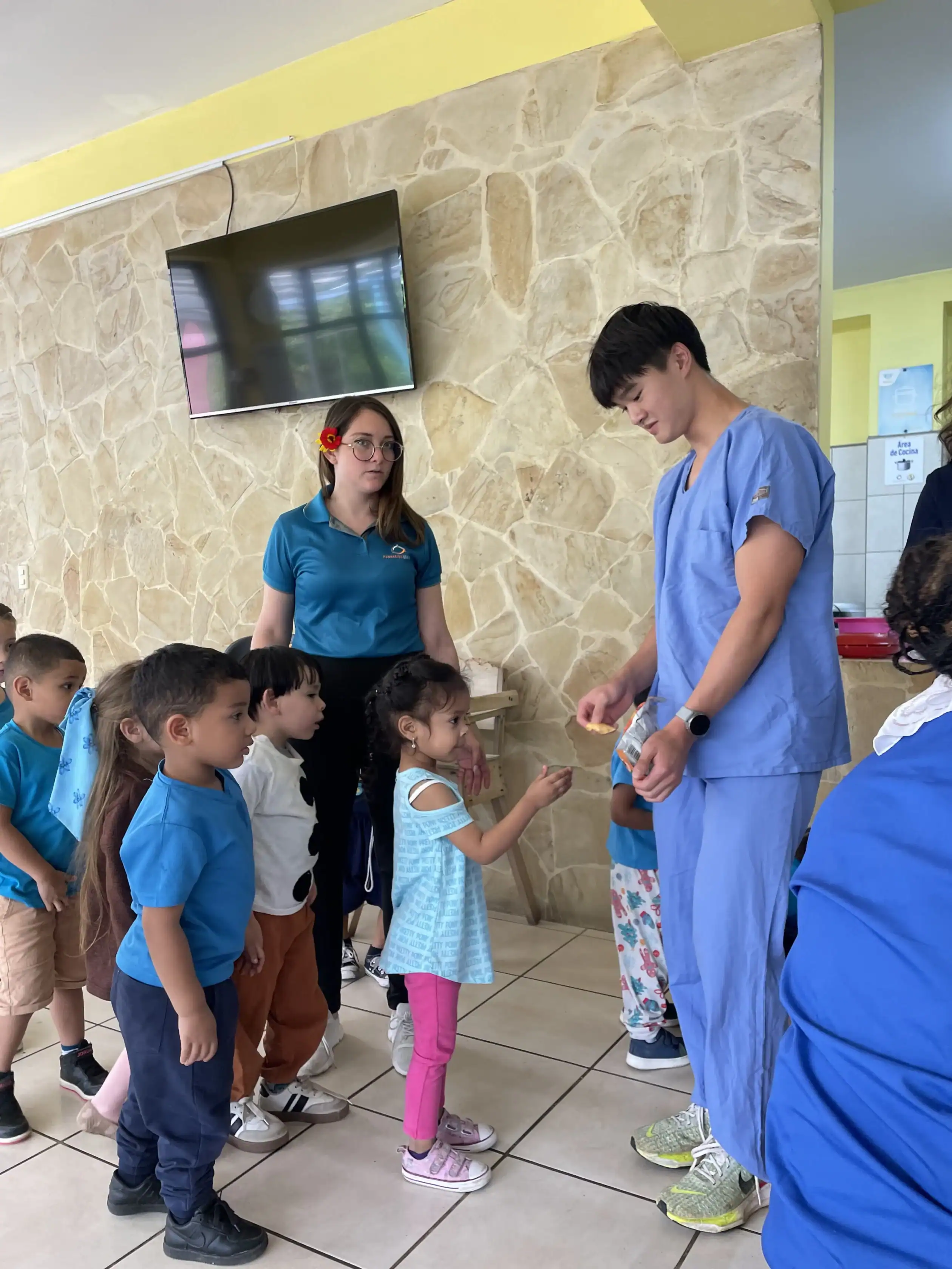 A man in medical scrubs hands out items to young children lined up indoors during a teen medical trip abroad, while a woman and other kids watch.