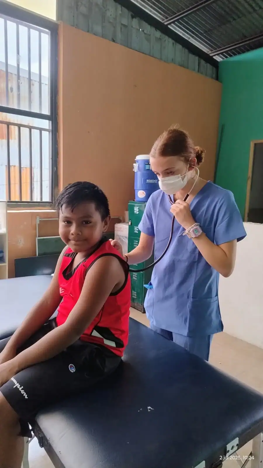 A healthcare worker in scrubs uses a stethoscope to examine a seated boy in a red shirt during a teen medical trip abroad at a local clinic.
