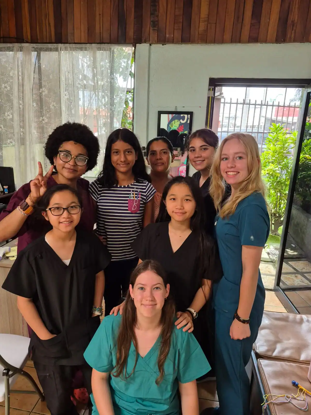 A group of eight young women, some wearing scrubs, pose and smile together indoors near a window with plants visible outside, capturing a moment from their teen medical trip abroad.